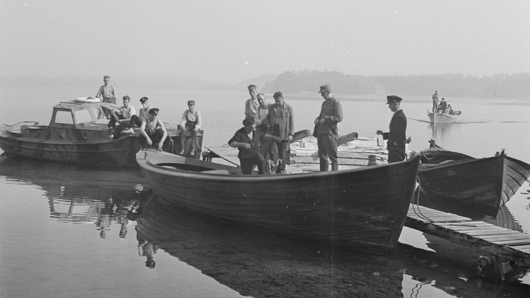 Flera personer står och sitter i roddbåtar och en motorbåt vid en brygga på en sjö. Texten ”SA-Kuva” syns i nedre högra hörnet.