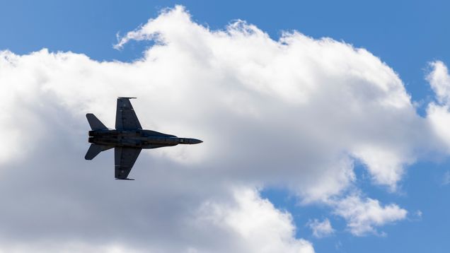 Silhouette of a fighter jet seen from below, flying in the air, with a large white cloud and blue sky in the background.