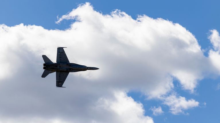 Silhouette of a fighter jet seen from below, flying in the air, with a large white cloud and blue sky in the background.