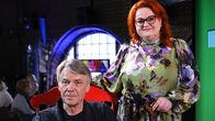 Harmaantunut mies istuu punaisessa pinnatuolissa. Hänen vieressään oikealla seisoo keski-ikäinen punatukkainen nainen.