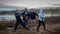 Neljän henkilön ryhmä seisoo ulkona joen rannalla sumuisessa maisemassa. Kukaan ei katso kameraan.