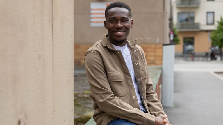 A smiling young African man sits on a wall in an urban setting.