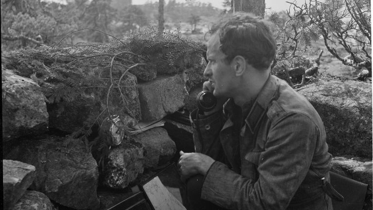 En soldat står vid en stenmur utomhus och håller en telefonlur; teknisk utrustning syns i förgrunden.