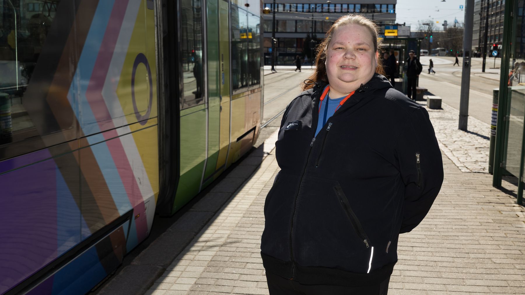 En kvinna står på en spårvagnshållplats bredvid en spårvagn i stadsmiljö.
