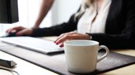 Kaffekopp i förgrunden, människa som skriver på dator i bakgrunden