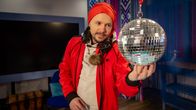Mies jouluisissa vaatteissa katselee diskopalloa. Kaulassa hänellä on headset-kuulokkeet. Taustalla näkyy iso televisio ja värikästä koristelua.