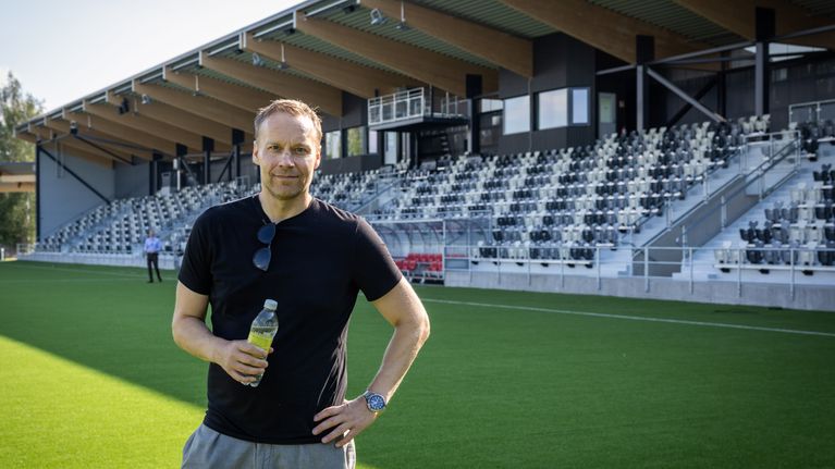 Mies seisoo jalkapallokentällä vesipullo kädessään. Taustalla on jalkapallostadionin katsomo. 