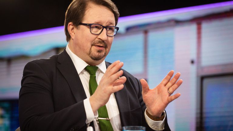 A man wearing a dark suit, green tie and glasses gestures as he speaks while seated in a TV studio.