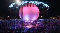 A full audience watches musicians on a theatre-in-the-round type stage with a large purple ball above the performers.