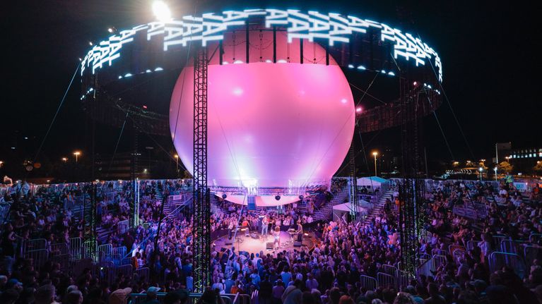 A full audience watches musicians on a theatre-in-the-round type stage with a large purple ball above the performers.