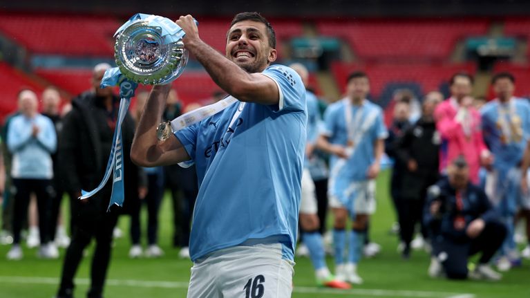En fotbollsspelare i Manchester Citys ljusblå matchtröja lyfter en pokal på en fotbollsplan, omgiven av lagkamrater.