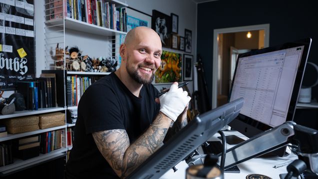 Henkilö istuu työpöydän ääressä digipiirtopöydän ja suuren näytön edessä, taustalla hyllyjä ja seinällä julisteita.