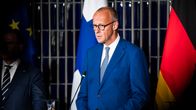 En man i blå kostym står vid en talarstol med mikrofon, framför EU:s, Finlands och Tysklands flaggor.