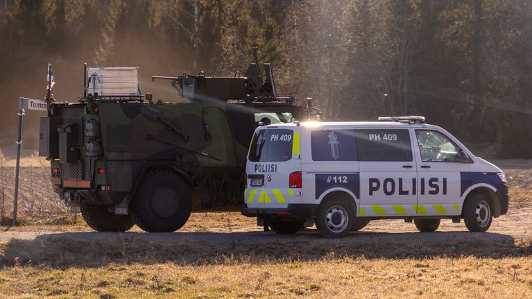 A green-and-black armoured vehicle and a blue-and-white police van parked on a sunny field with trees in the background.