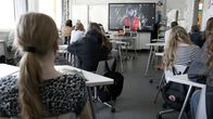 Kuvassa on koululuokka, jossa oppilaat istuvat pulpeteissa ja katsovat luokan etuosan videotaulua seinällä.