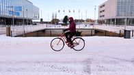 Person wearing a dark red winter coat riding a bicycle on a snowy city street, with two concrete and glass buildings seen in the background.