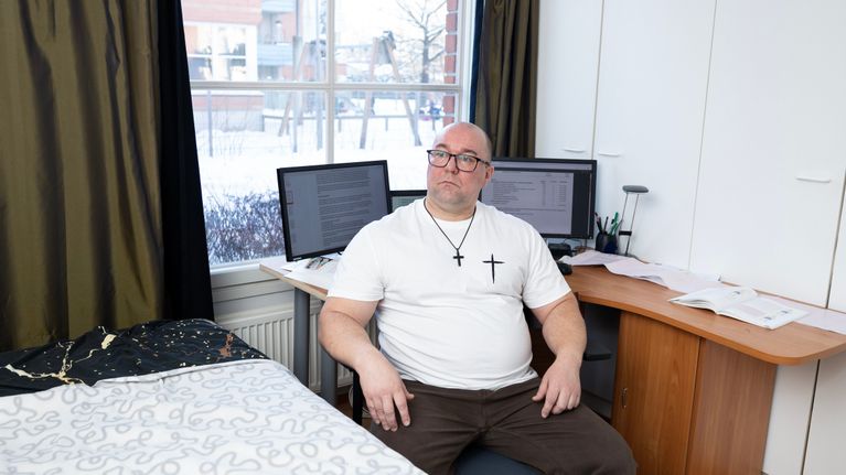 Mies istuu työpöydän ääreessä, taustalla tietokoneita.