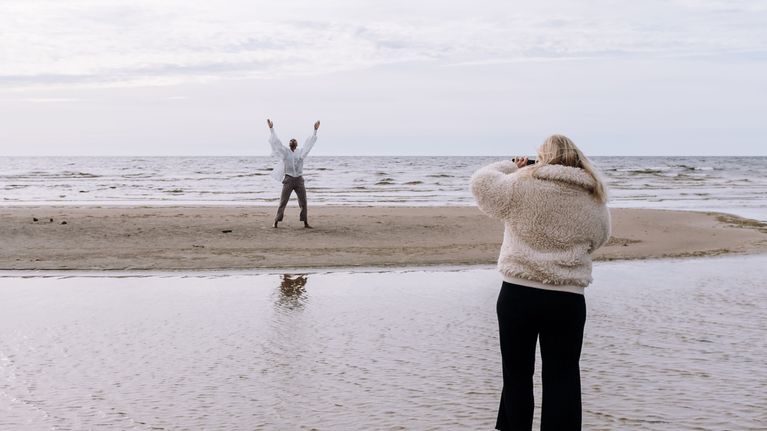 Emilia Hernesniemi ja Jani Toivola Hailuodossa meren rannalla. Hernesniemi kuvaa Toivolaa, joka tanssii hiekkarannalla valkoisessa paidassa.