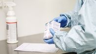 A healthcare worker in protective clothing and gloves holds two medicine packages at a stainless-steel workbench.