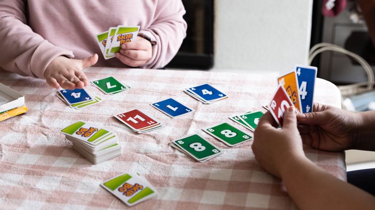 Kaksi henkilöä pelaa pöydän ääressä Skip-Bo-korttipeliä ruutukuosisella pöytäliinalla.