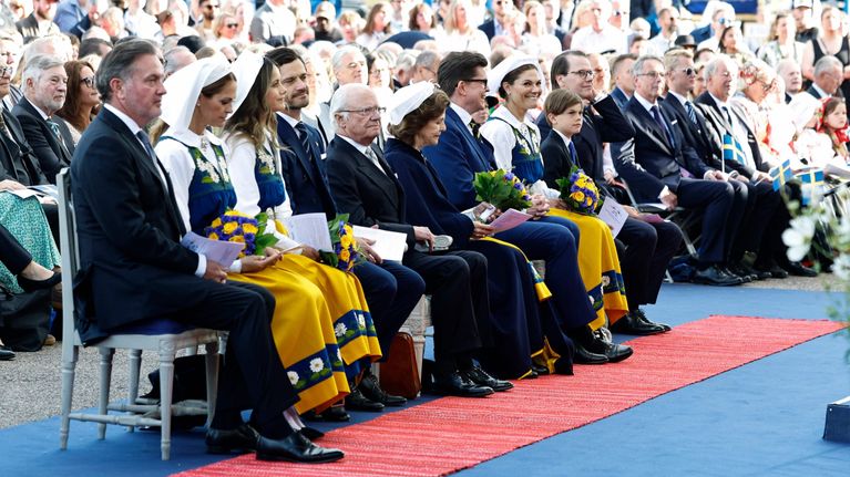 Svenska kungafamiljen sitter på första raden i publiken. Prinsessorna ahr folkdräkt och blombuketter i händerna.