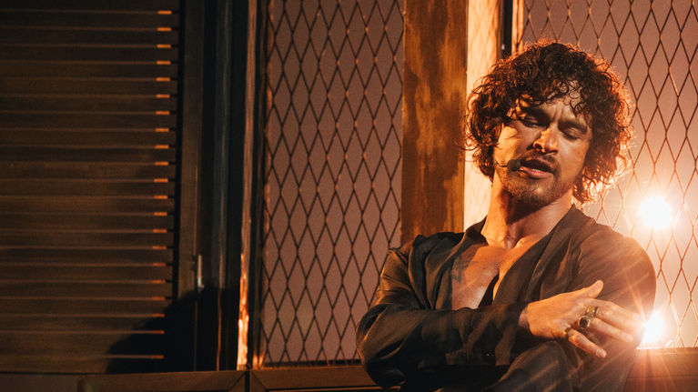 Man with dark hair sitting on a bench singing on a stage with dramatic lighting.