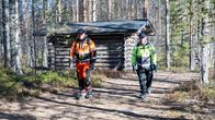 Kaksi miestä työvaatteissa kävelee metsätiellä pois pyöröhirsisen halkovajan luota.