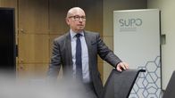 Silmälasipäinen kalju mies seisoo neuvotteluhuoneessa ja nojaa tuolin selkään.