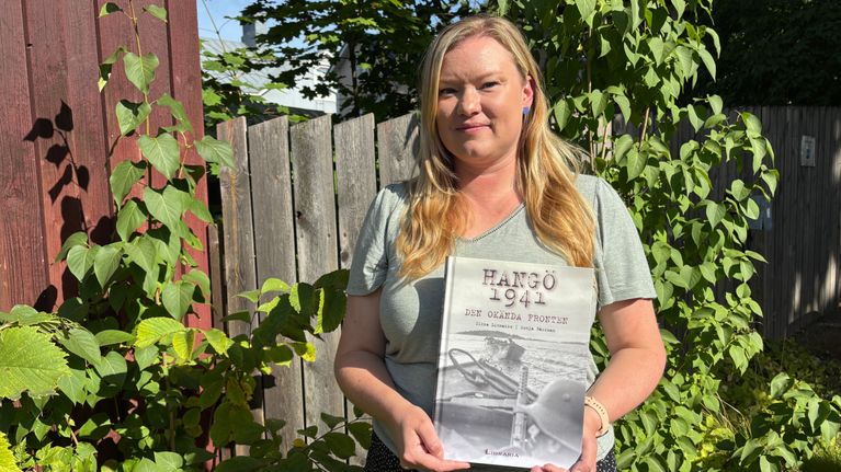 En kvinna står utomhus framför ett trästaket och håller i en bok med titeln "Hangö 1941 – Den glömda fronten".