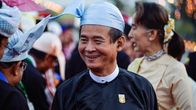 Win Myint hymyilee perinteisessä myanmarilaisessa puvussa, vaaleansininen gaung baung -päähine ja tumma taik-pon eingyi -takki.
