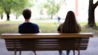 Två personer sitter på en parkbänk med ryggen mot kameran. Det är en solig och grön dag. Bilden är suddig.