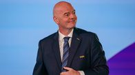 Pukuun ja solmioon pukeutunut mies seisoo sinertävän taustan edessä, puvuntakissa lukee ”FIFA”.