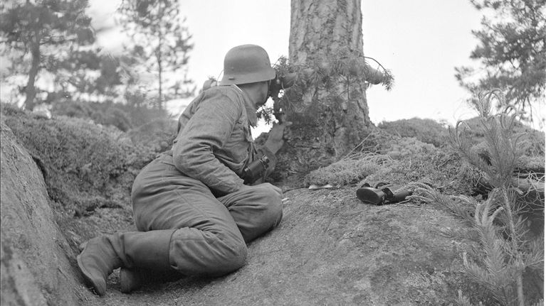 En soldat i uniform och hjälm sitter på knä bakom en tall, håller ett gevär och spanar framåt. Texten "SA-Kuva" syns i hörnet.