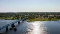 Aerial view of a blue steel bridge over a river with sun glinting on it and trees and buildings on the opposite shore.