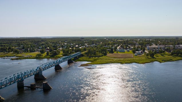 Aerial view of a blue steel bridge over a river with sun glinting on it and trees and buildings on the opposite shore.