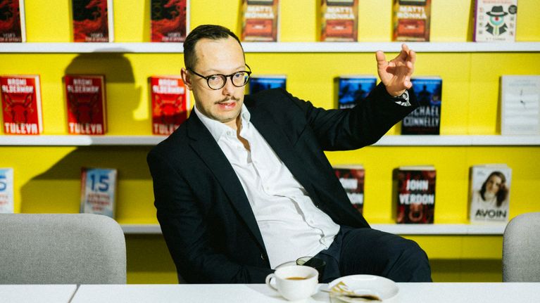 Henkilö istuu pöydän äärellä. Taustalla seinällä on useita kirjahyllyjä, joilla on kirjoja.