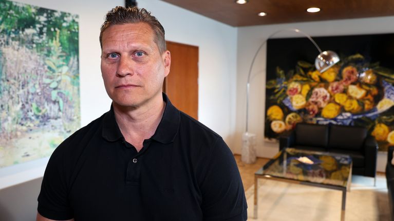 Tuomas Heikkinen, en person i svart tröja står i ett rum med tavlor på väggarna och en soffa framför ett glasbord.