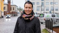 På bilden ser vi en kvinna iklädd en svart, vadderad jacka och en färgglad halsduk som står på en kullerstensgata en molnig dag.