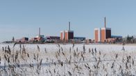 A row of large red and white industrial buildings seen across a snow-covered frozen bay with reeds in the foreground.