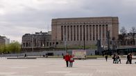 Finnish Parliament building in downtown Helsinki. People are walking on a square, with a cloudy, grey sky in the background.