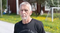 En man står på en landsväg framför ett gammalt rött hus med vita knutar. Marken är täckt av gröna växter och gula blommor.