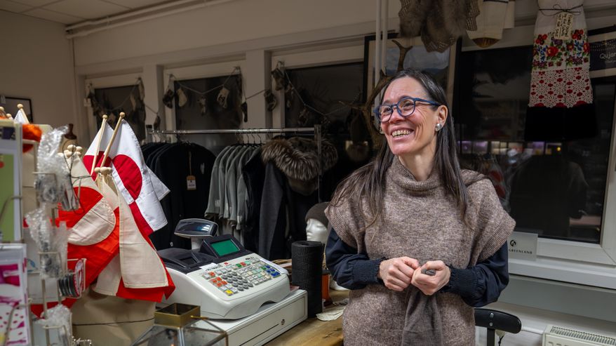 Tumma, pitkähiuksinen nainen, Vivi Schultz, puodissaan kassan takana. Neoulos kädessään. Hymyilee.