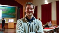 Valokuva nuoresta miehestä seisomassa sisätiloissa, jossa on punaiset verhot ja ihmisiä pöytien ääressä.