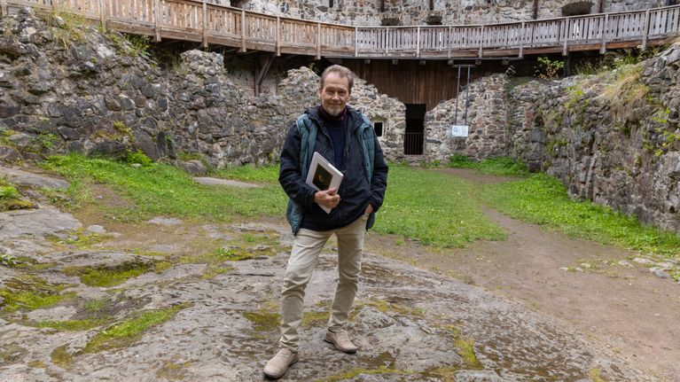 En person står på en stenig innergård framför en gammal stenmursbyggnad och håller i en bok.