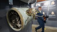 Etualalla roikkuu katosta rikkinäinen ja kolhiintunut metallinen leveä putki, jonka kyljestä törröttää johtoja. Taustalla kävelee hahmo puvussa.