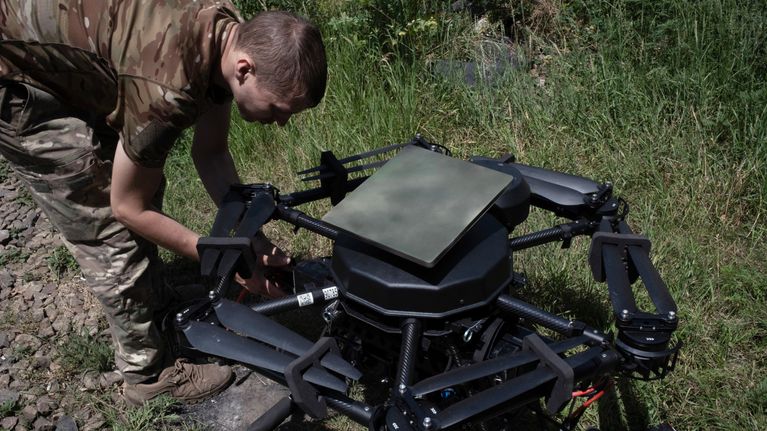 Ukrainalainen sotilas työskentelee suuren mustan droonin parissa ulkona nurmialueella. 