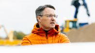 Mies oranssissa takissa seisoo ulkona työmaakoneiden ja hiekkakasan edessä. 