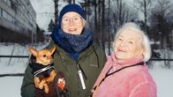 Kaksi henkilöä talvisessa puistossa poseeraavt iloisesti hymyillen kameralle. Nuoremmalla henkilöllä o pieni rottakoira sylissään. 