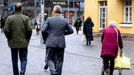 Vanhus rollaattorin kanssa ja kaksi pukuun pukeutunutta henkilö kadulla.
