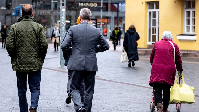 Vanhus rollaattorin kanssa ja kaksi pukuun pukeutunutta henkilö kadulla.